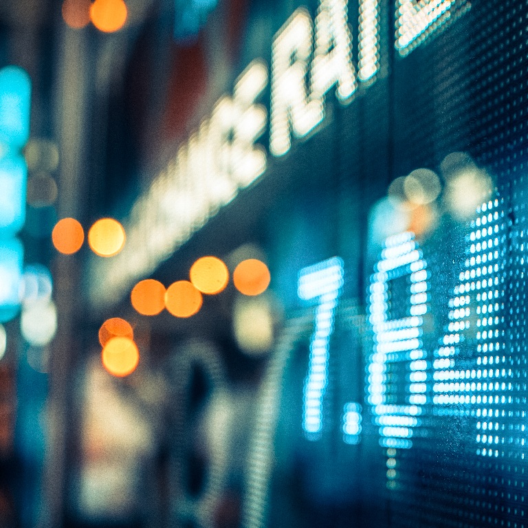 Close-up of a blue LED market ticker showing numeric quotes against a blurred city street background, conveying financial trading data.